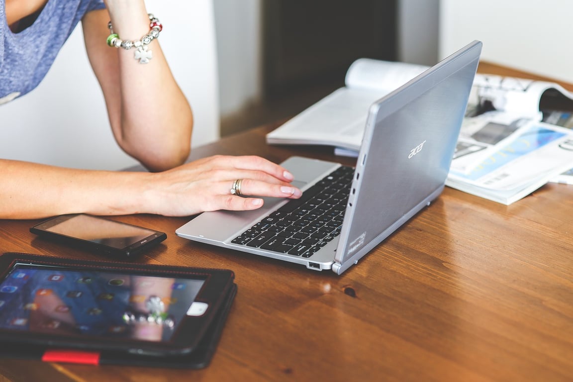 A woman working on a laptop, with a few magazines, a phone, and an iPad on the table.